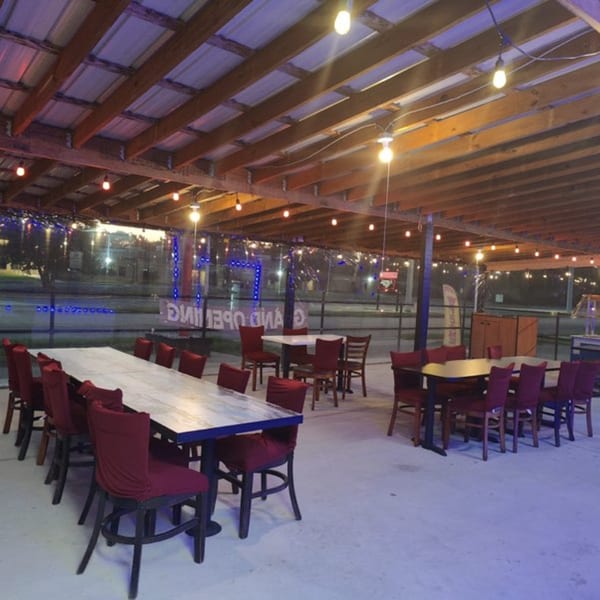 Outdoor dining area with long tables, red chairs, string lights, and a "Grand Opening" sign.