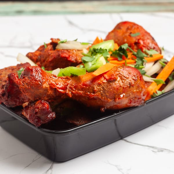 Indian 5 Stars Restaurant's chicken drumsticks, marinated in red spices, garnished with fresh vegetables in a black container.